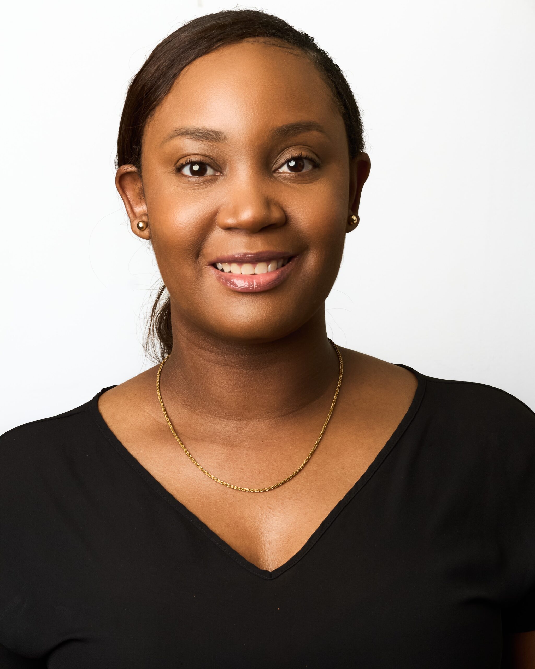 Close-up portrait of a smiling Black woman wearing a black V-neck top and a thin gold necklace against a white background.
