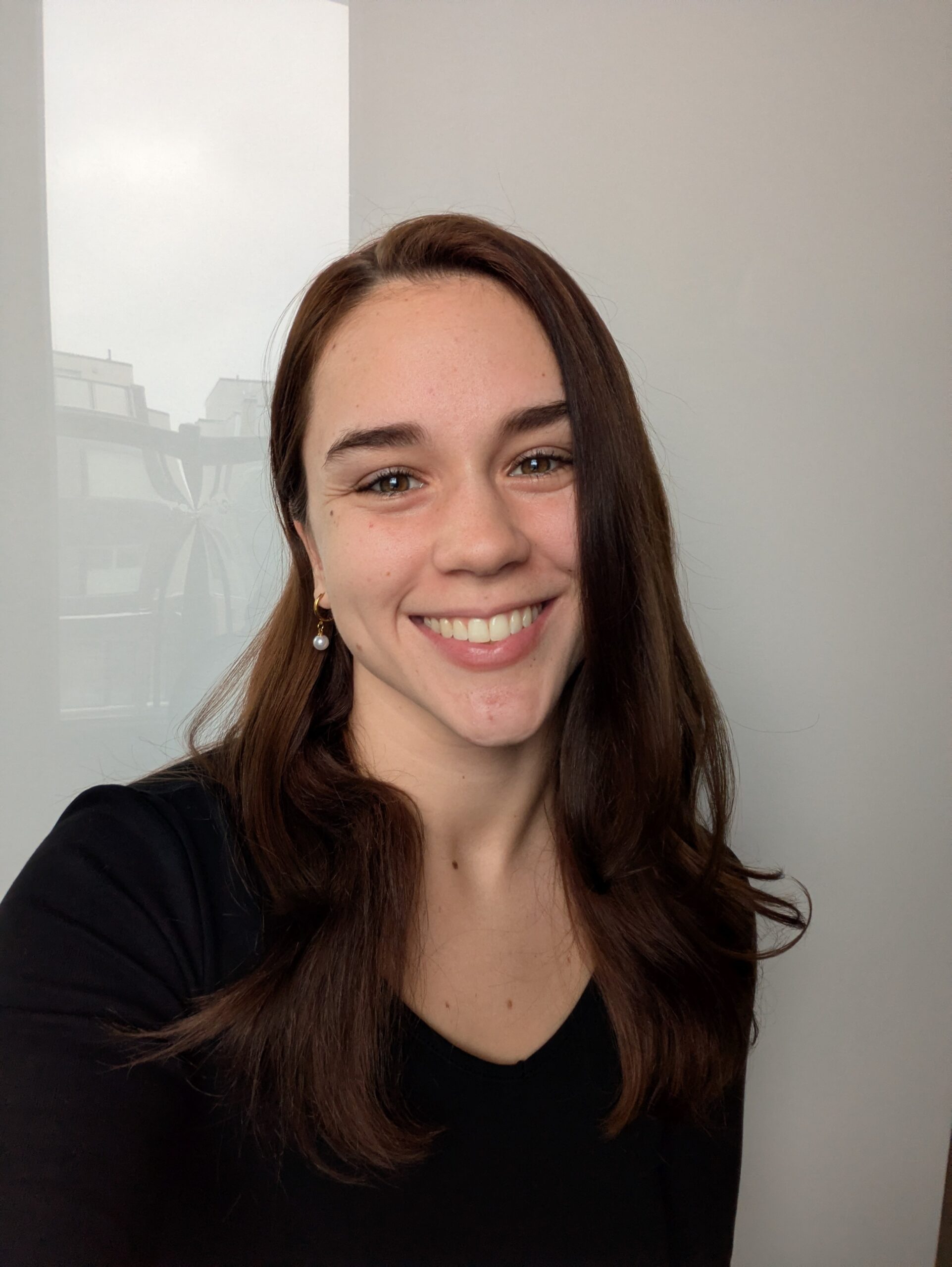 Smiling young woman with long brown hair taking a selfie indoors, wearing a black top.