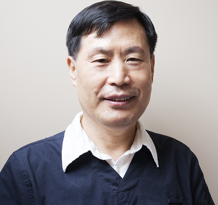 Portrait of a middle-aged Asian man smiling at the camera, wearing a dark navy shirt over a white collared shirt against a neutral background.